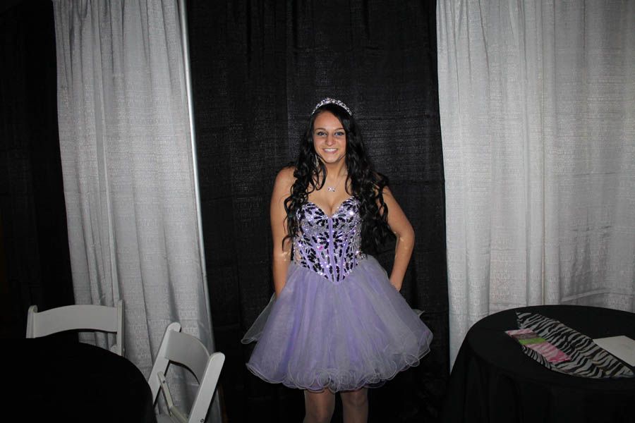Woman in a purple dress and tiara smiling, posing in front of curtains and a table.