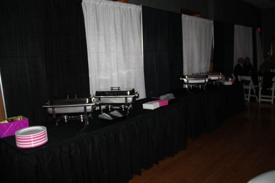 Buffet with black tablecloths, white and pink plates, and food warmers. Black and white curtains in the background.