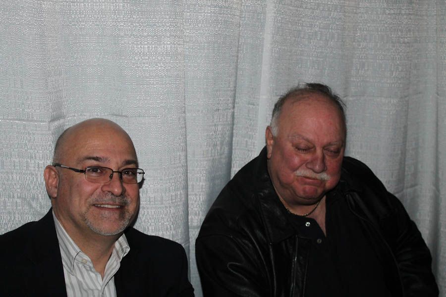 Two men seated, smiling; one bald with glasses, other with mustache, black jacket.