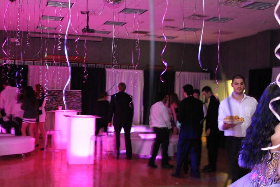 Party scene with people, streamers, and pink lighting in a large room. Some people are standing, others are mingling.