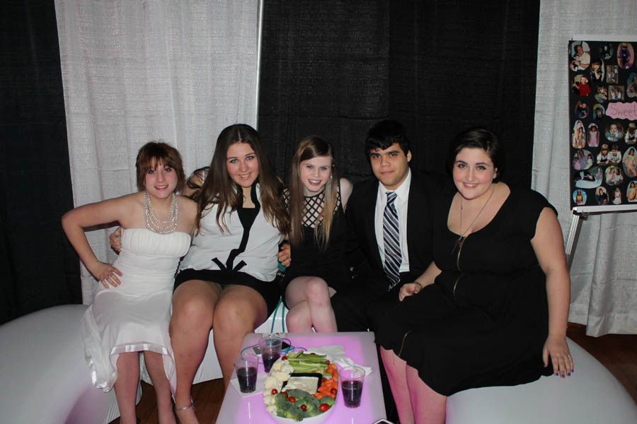 Group of five people seated on a couch; formal attire, smiling. A small table with snacks and drinks is in front of them.