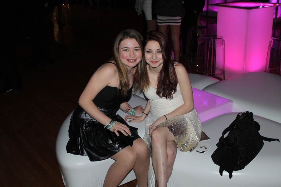 Two young women in party dresses sitting on white sofa; illuminated background.