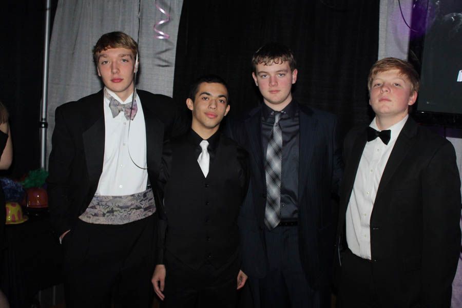 Four young men in formal attire, posing together at an event with dark background.