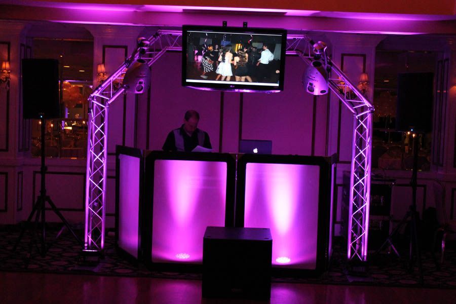 DJ setup at an event, with purple stage lighting, a screen, and sound equipment. DJ is at a laptop.