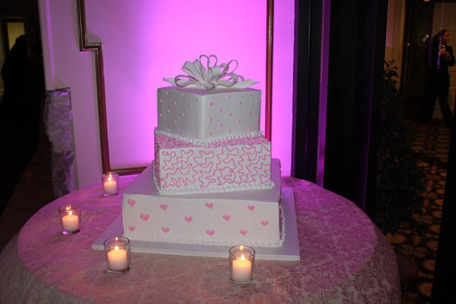 Three-tiered square cake decorated with pink hearts, bows, on a round table, lit candles, purple lighting.