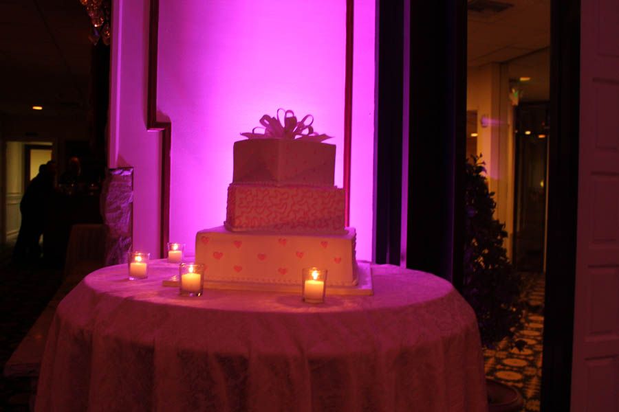 Three-tiered cake with pink hearts and bow on a table lit by candles, bathed in pink light.