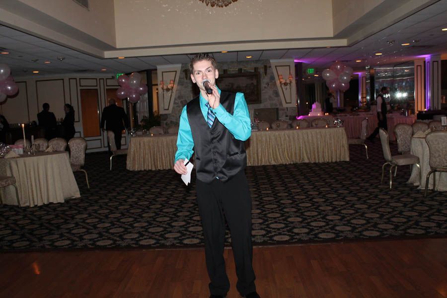 Man in turquoise shirt and black vest speaks into microphone at an event with tables and decorations.