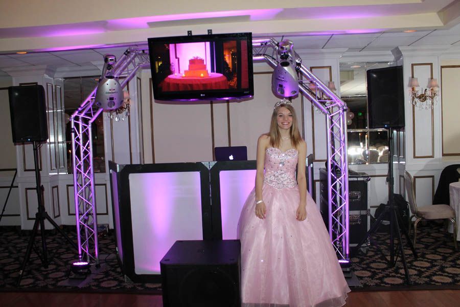 Girl in pink gown stands at a DJ setup with purple lighting, a TV, and speakers in a ballroom.