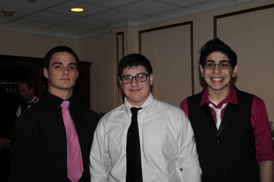 Three young men in formal attire, smiling at the camera indoors. One in pink tie, one in black tie, and one in vest.
