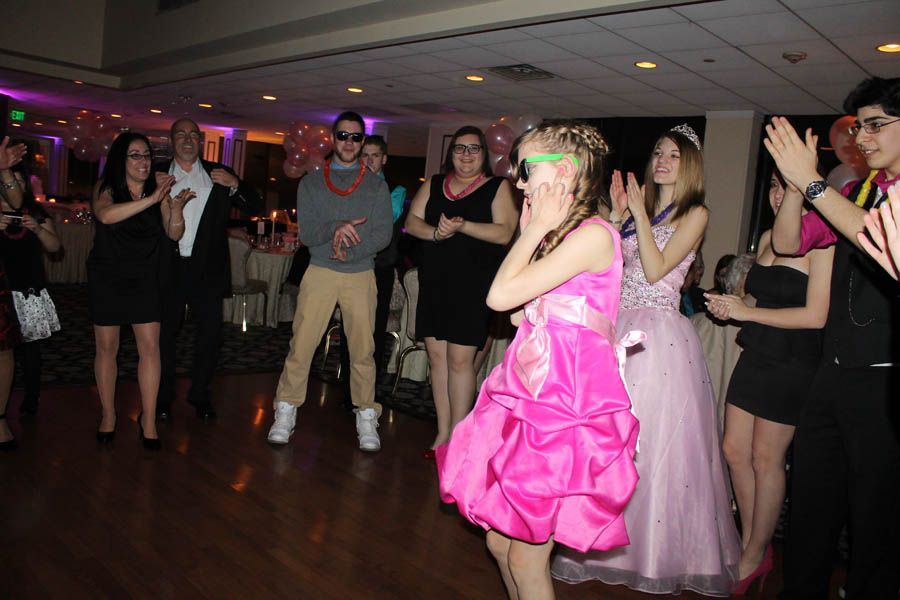People clapping for a girl in a pink dress, dancing in a room with balloons and tables.