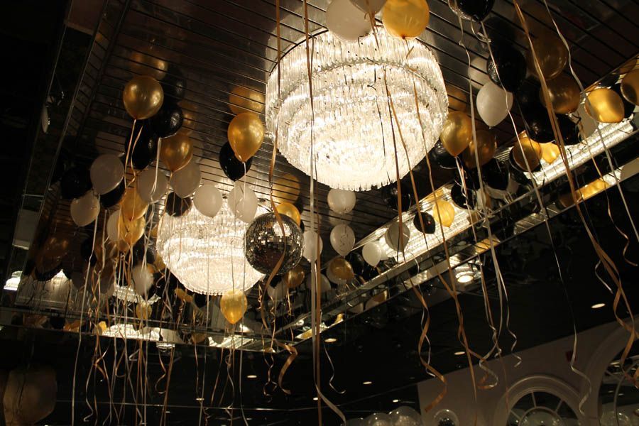 Gold, black, and white balloons decorate a room with large chandeliers and mirrored ceiling.