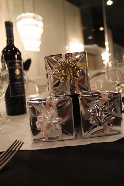 Small, silver-wrapped gift boxes with bows stacked on a table set for dinner; wine bottle in background.