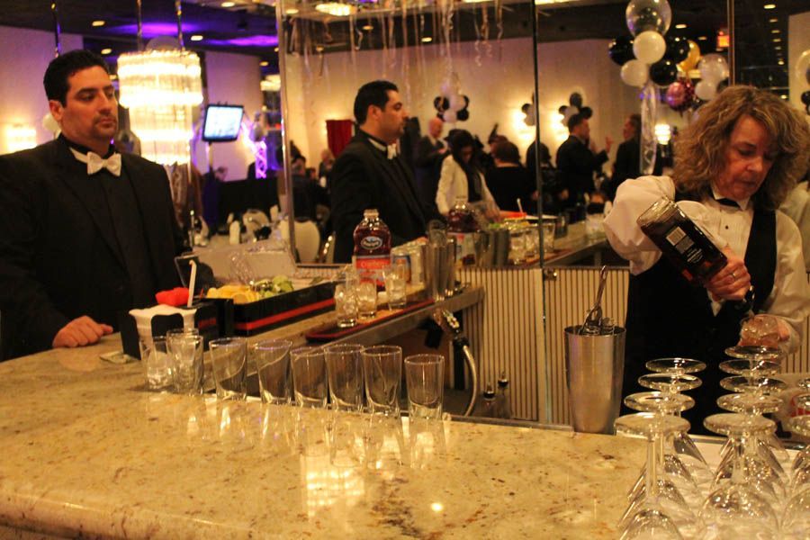 Two bartenders in tuxedos behind a marble bar, one pouring a drink. Balloons and guests in the background.