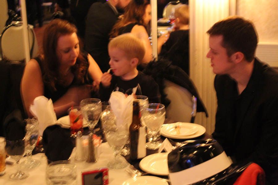 Three people at a dinner table: woman, boy eating, and man talking. Table set with glasses and white cloth.