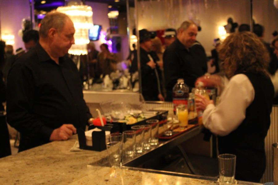 Bartender pouring a drink at a crowded bar. Man in black shirt waits at the counter. Dimly lit, indoor event.