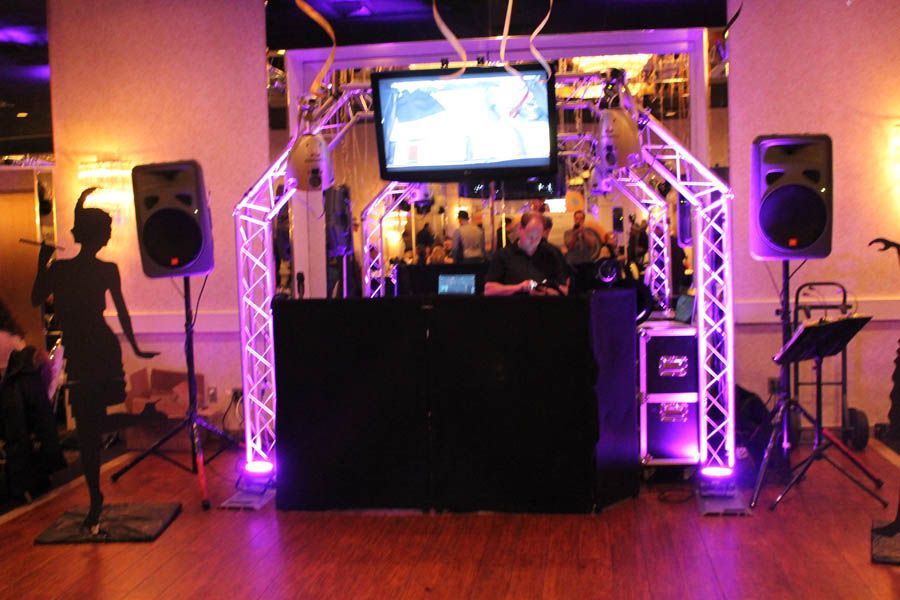 DJ setup with speakers, lights, screen, and a person behind a black table in a room.