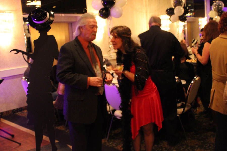 Man and woman in evening wear chatting at a party. Decorative silhouette, balloons, and other guests visible.