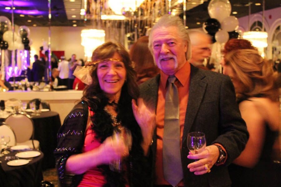 Man and woman smiling at an event, man holding a glass of wine, both in party attire.