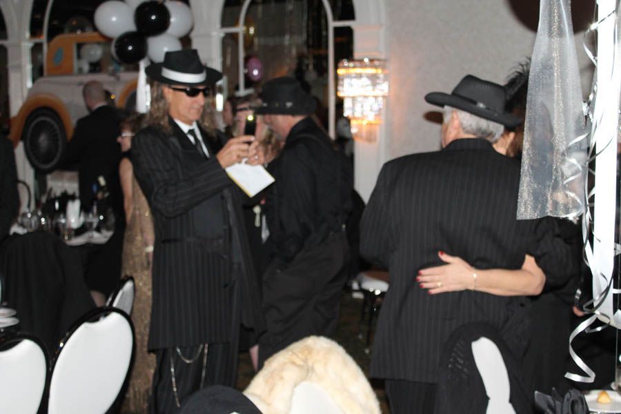 People in pinstripe suits and hats at a party, toasting drinks. Decorative black and white balloons and a car.