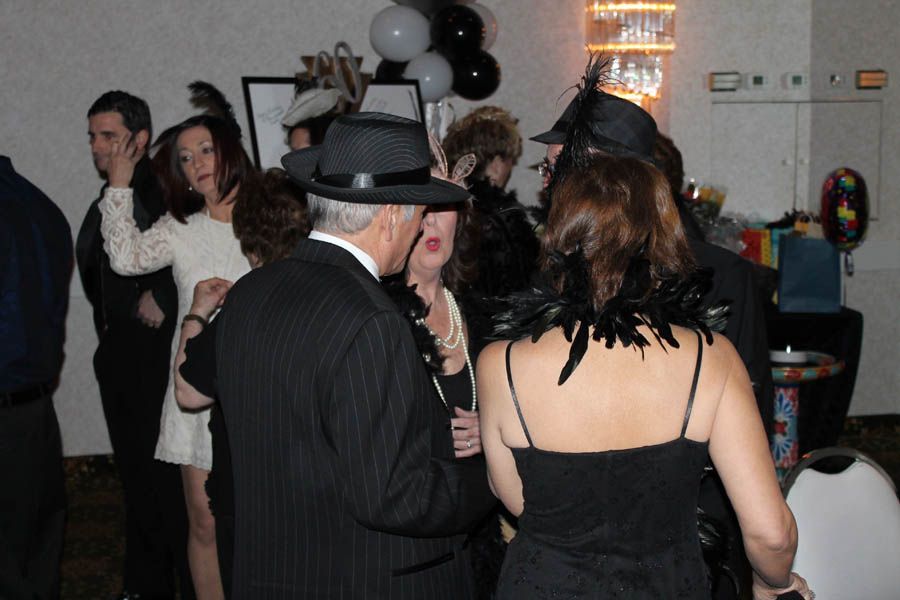 People in a room, some wearing hats and feather boas, possibly at a party. Black and white balloons in background.
