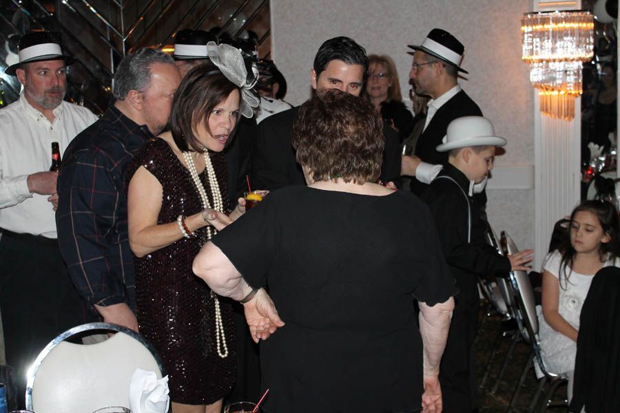 Group of people in 1920s attire at an event, some wearing hats and holding drinks, indoors.