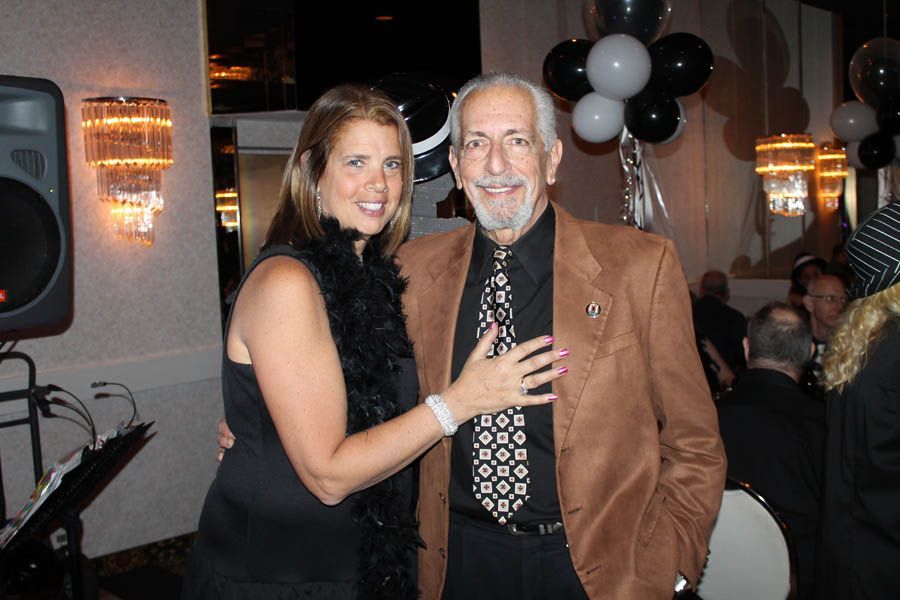 Woman with black dress and boa, hand on man's chest. Man in brown blazer, patterned tie. Party setting, balloons.