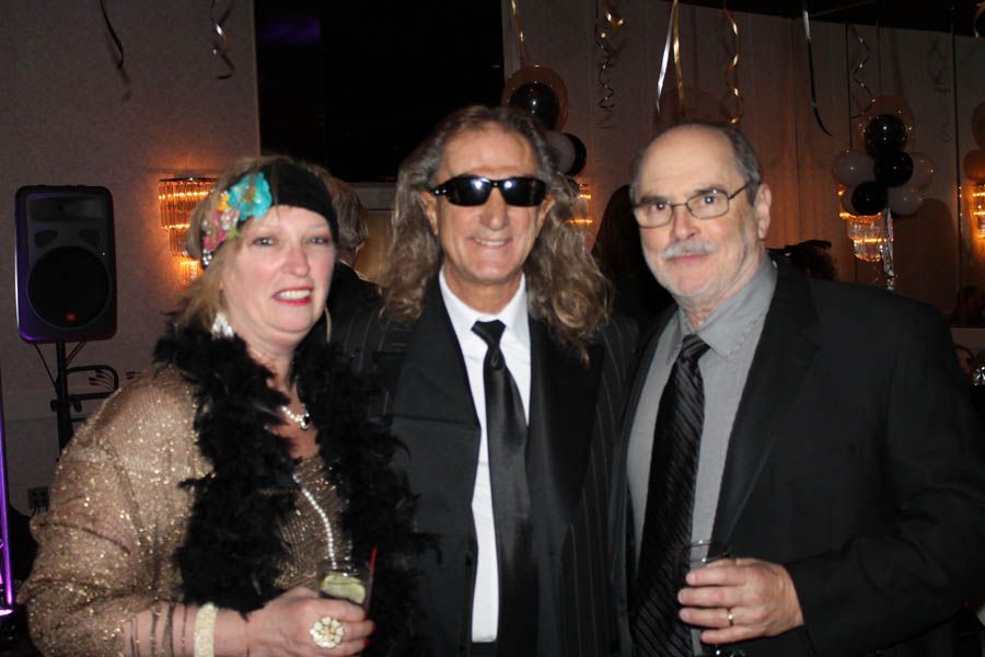 Three people in formal attire pose for a photo at a party. One wears a flapper headband and boa.