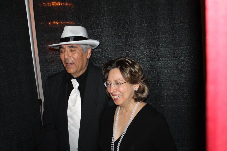 Man in fedora and suit with woman in black smiling, standing by a dark curtain.