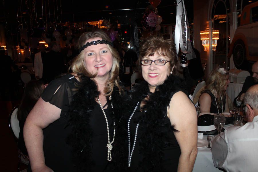 Two women in flapper attire at a party, wearing boas, beads, and headbands, smiling.