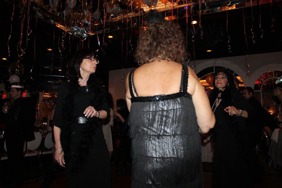 Three women in black dresses at a party, one dancing. Metallic decorations overhead. Dimly lit room.