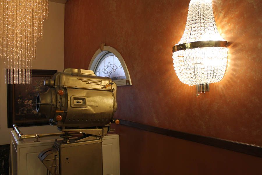 Vintage gold-toned film projector atop a cabinet, beneath a crystal chandelier and a sconce on a reddish-brown wall.