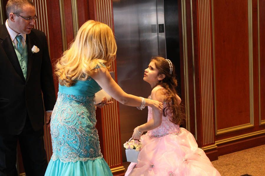 Woman in blue dress comforts girl in pink dress near elevator. Man in suit watches.