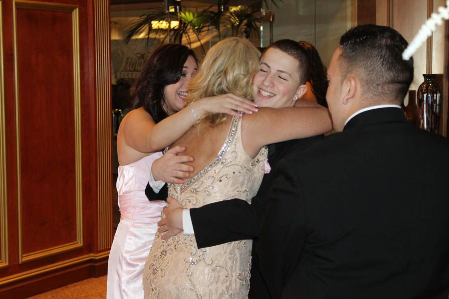 Group embraces, formal attire in a well-lit interior. A woman in a sparkling dress is embraced by three others.