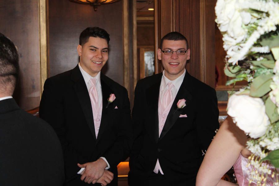 Two men in tuxedos, pink ties, smiling, in a decorated room, possibly at a wedding.