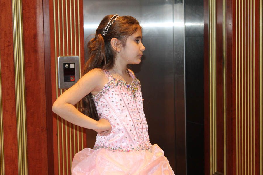 Girl in a pink formal dress, standing in front of an elevator in a wood-paneled room.