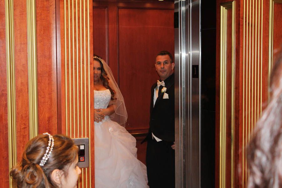 Bride and groom inside an elevator, bride in white gown, groom in tuxedo, looking at camera.