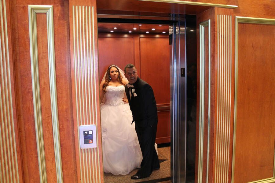 Bride and groom exit a wood-paneled elevator. The bride wears a white wedding dress, the groom a black suit.