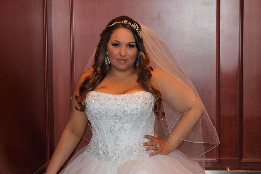 Bride in a strapless white wedding dress with beading, veil, and tiara, posing indoors with hand on hip.