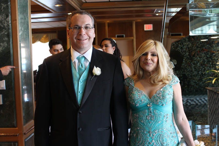 Couple in formal attire, smiling, walking indoors. Man in black suit, teal tie. Woman in teal dress, blonde hair.