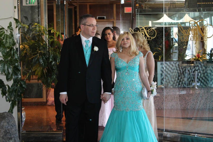 Couple in formal attire, holding hands, entering a building with glass doors, trailed by bridesmaids.