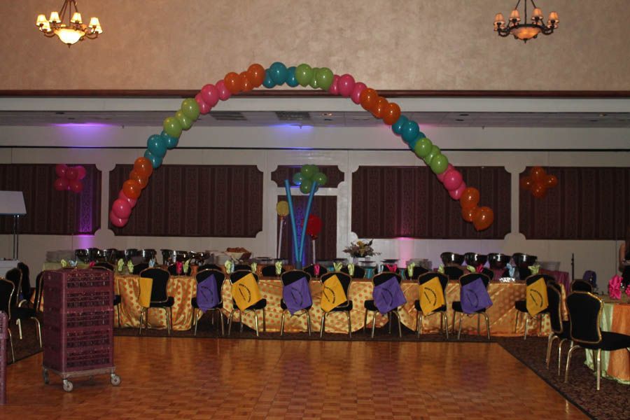 Balloon arch over a long banquet table with colorful decorations, in a ballroom.