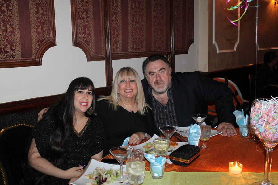 Three people smiling at a table in a restaurant. Woman with long dark hair on the left, two others smiling.