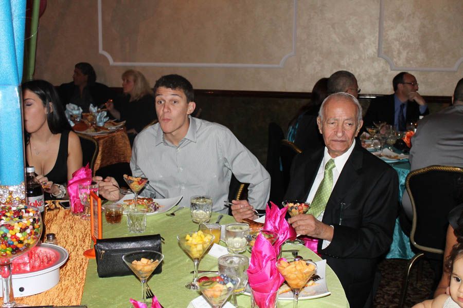 People at a round table with food and drinks. Man in gray shirt makes a face, next to a man in a suit.
