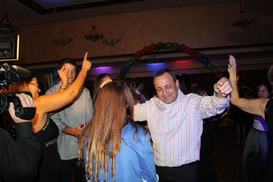 People dancing in a party setting; man in striped shirt with arms raised; archway of balloons.