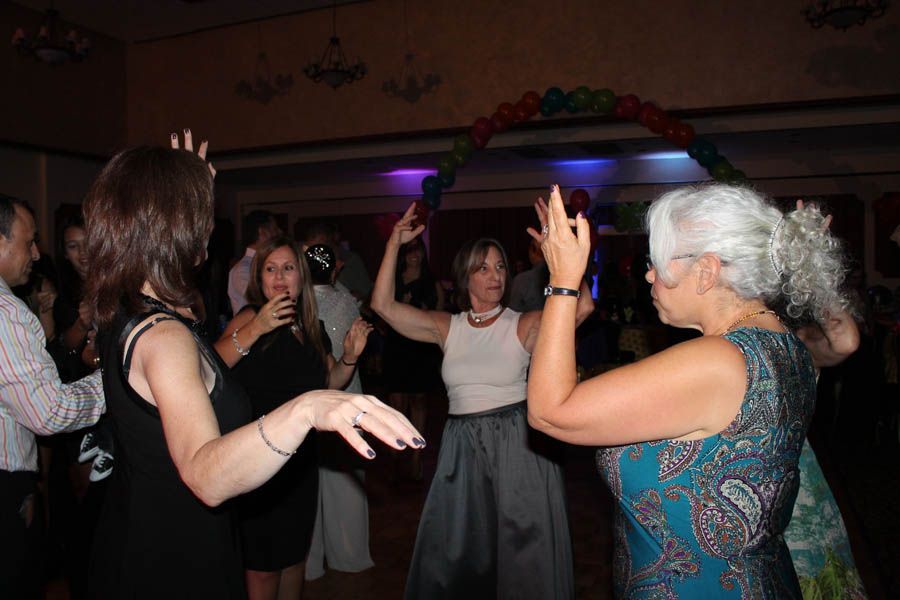 People dancing at a party under a balloon arch, some with arms raised. Dim lighting, formal wear.