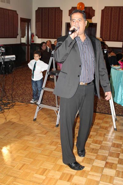 Man singing into a microphone, wearing a suit, on a dance floor. A child stands on a small ladder nearby.