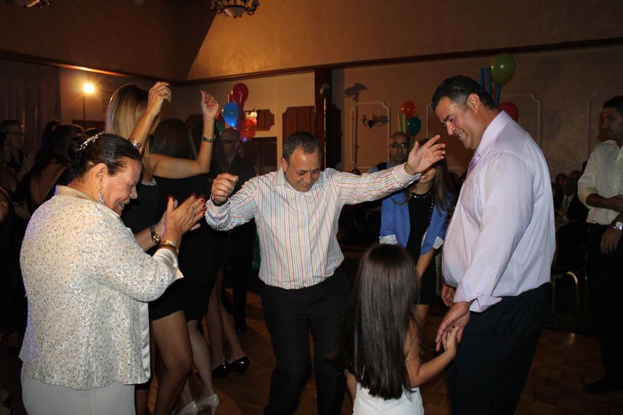 People dancing at a party in a decorated room. Man in center dances with arms out, girl holding hands. Others clap.