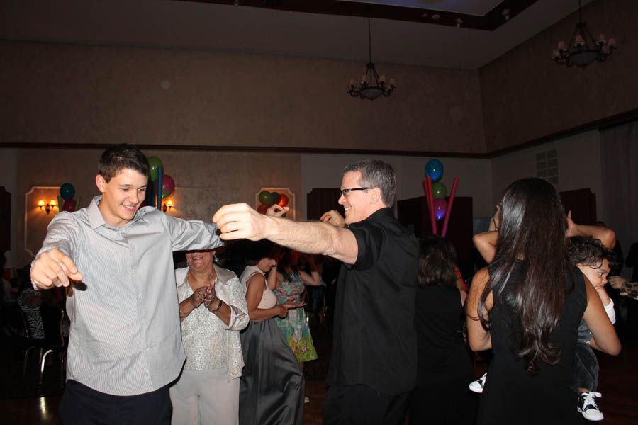 People dancing at an event, men in the foreground, woman holding a child. Balloons in the background.