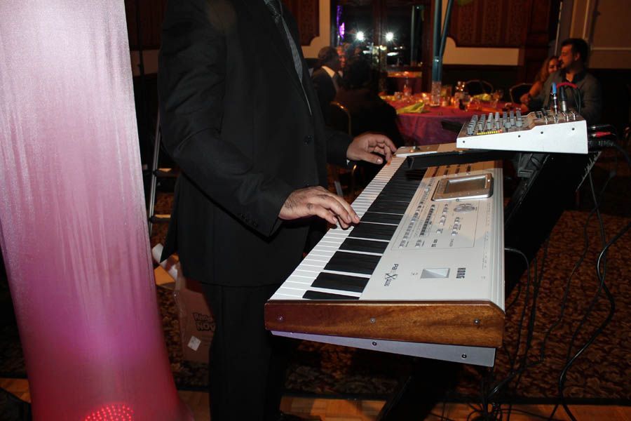 Person in a suit playing a keyboard on a stand. Stage setting with sound equipment and pink fabric.