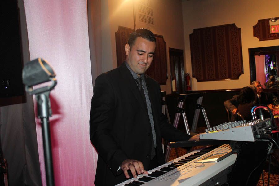 Man in suit playing keyboard, smiling in a dimly lit venue with a microphone and sound mixer.